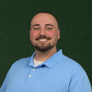 Professional headshot of Nic Rubolino, the founder of Noble Lion Marketing with a friendly smile, and short brown hair, wearing a blue shirt against a green background.